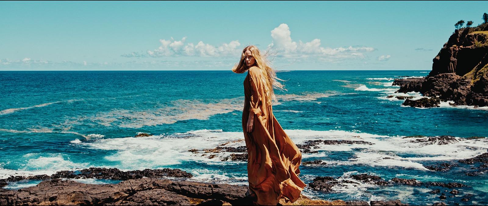 A woman in a flowing copper dress looking back across a rocky coastline