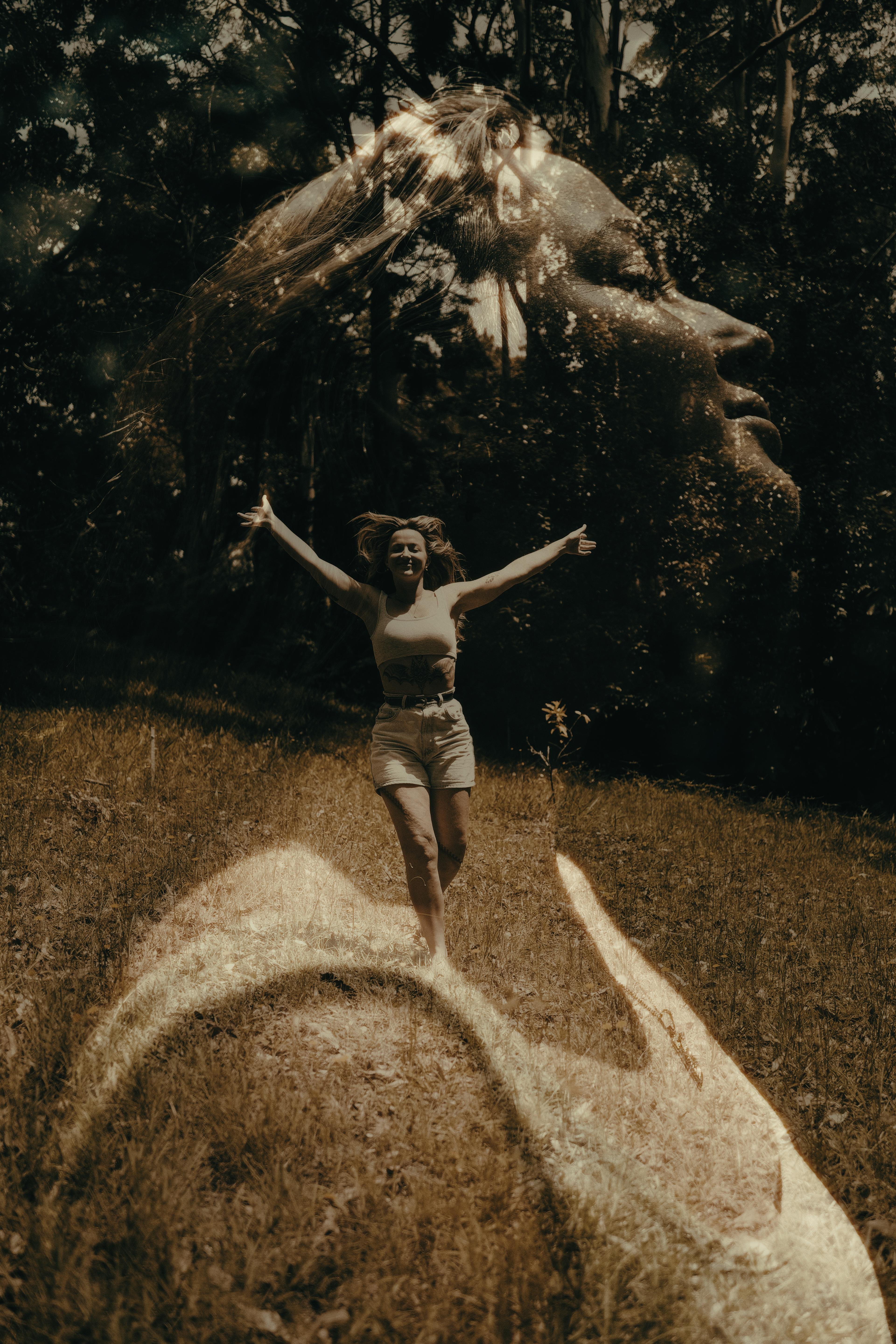 Double exposure of a woman jumping with arms outstretched in a field, superimposed with a large ghostly profile of a face made from the trees and light behind her