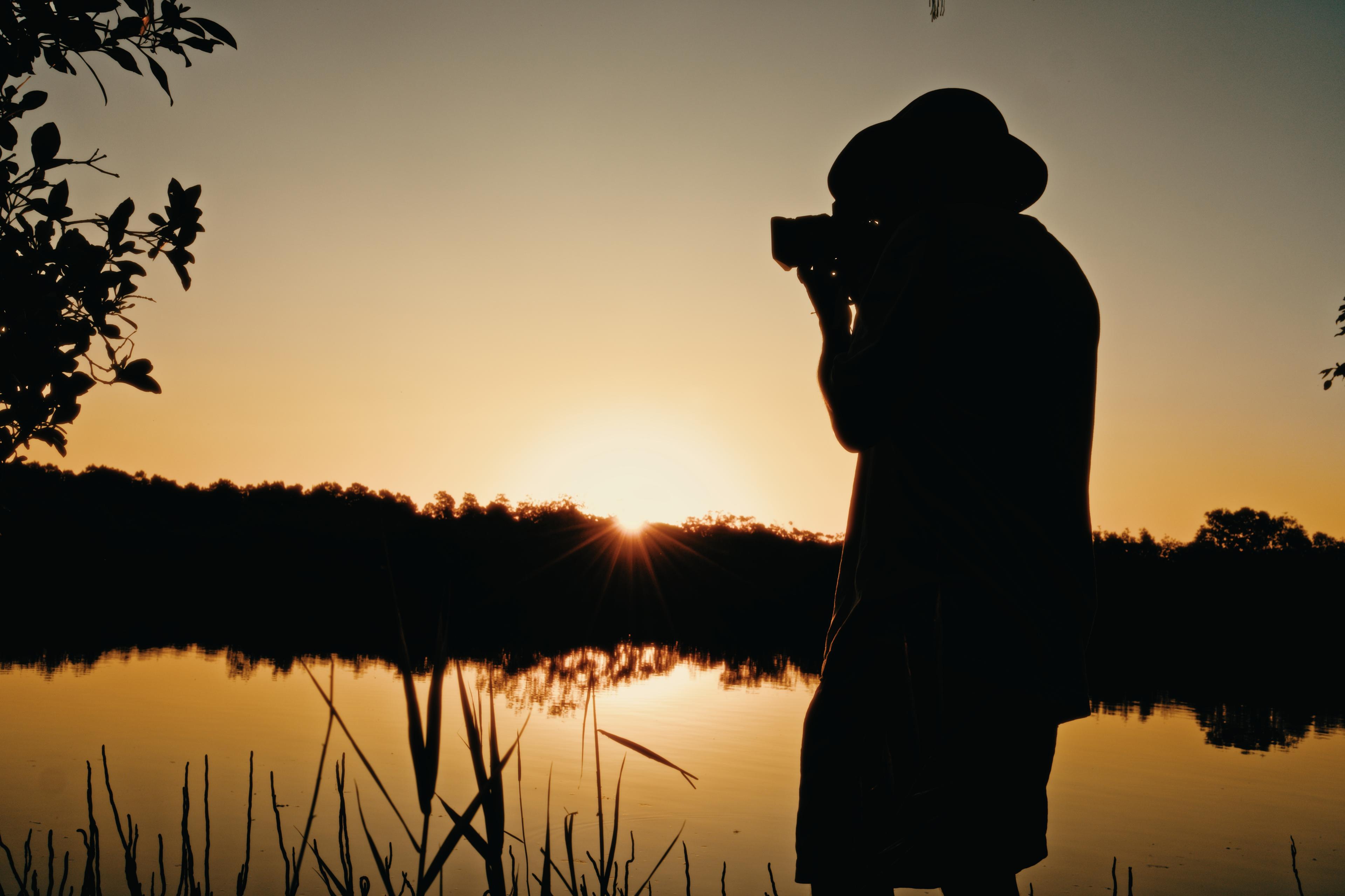 Silhouette of a photographer in a wide-brimmed hat, camera raised, sun bursting through the trees behind a still lake