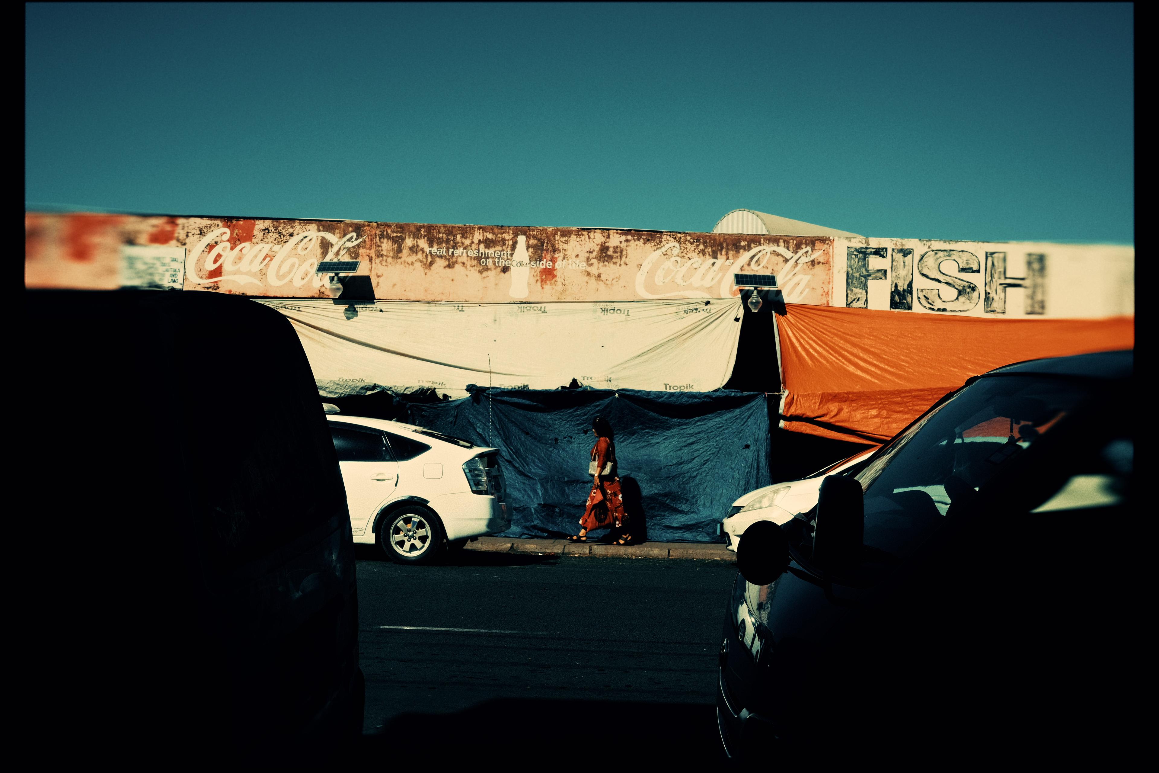 A woman in a red dress walking past a wall painted with old Coca-Cola and FISH signage, tarpaulins in orange and blue, two white cars parked in front