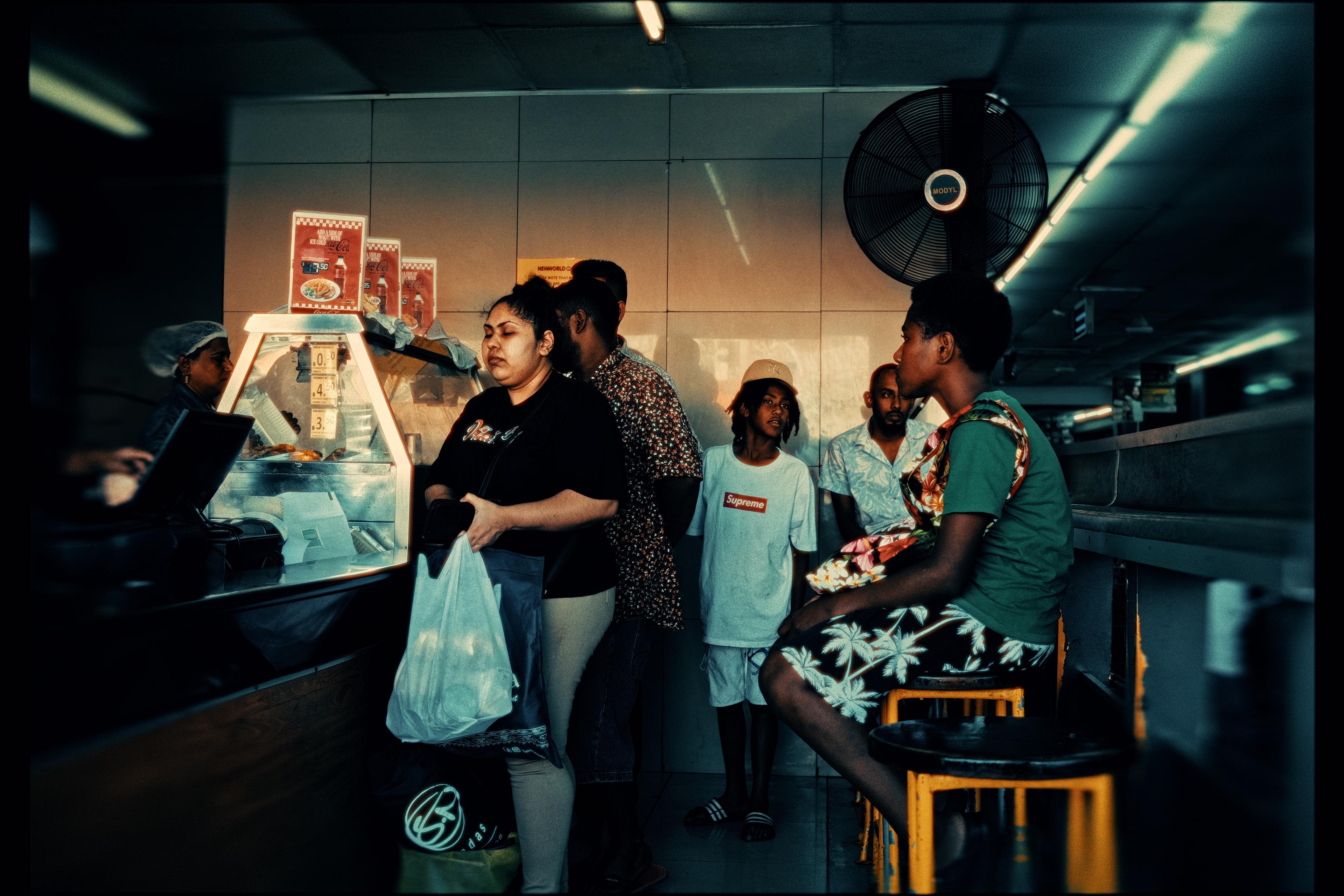 A queue of people waiting inside a takeaway shop, warm light and a wall fan behind them