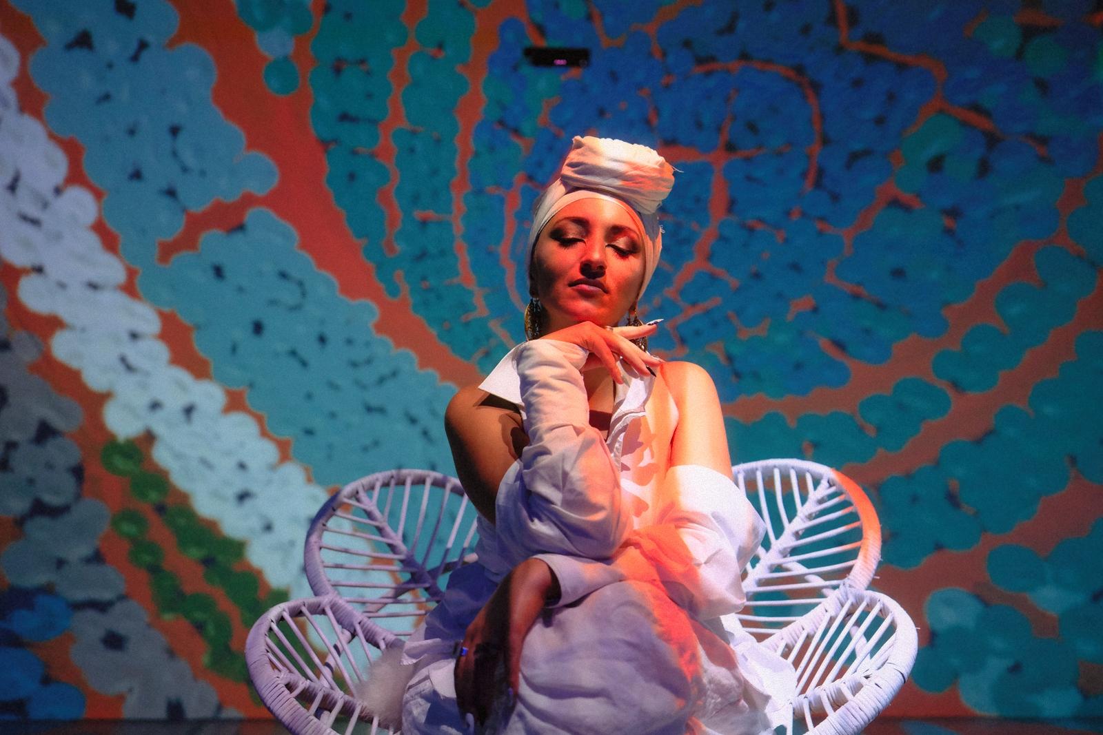 Juman seated in a rattan chair against a projected blue and orange backdrop, closer crop