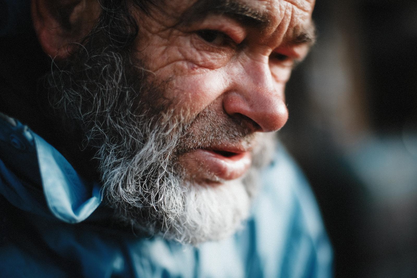 Close portrait of a bearded man in a blue jacket, lit from the side, looking down