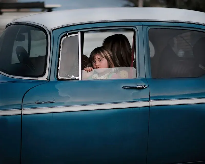 A young girl looking out from the window of a vintage blue car, an adult beside her