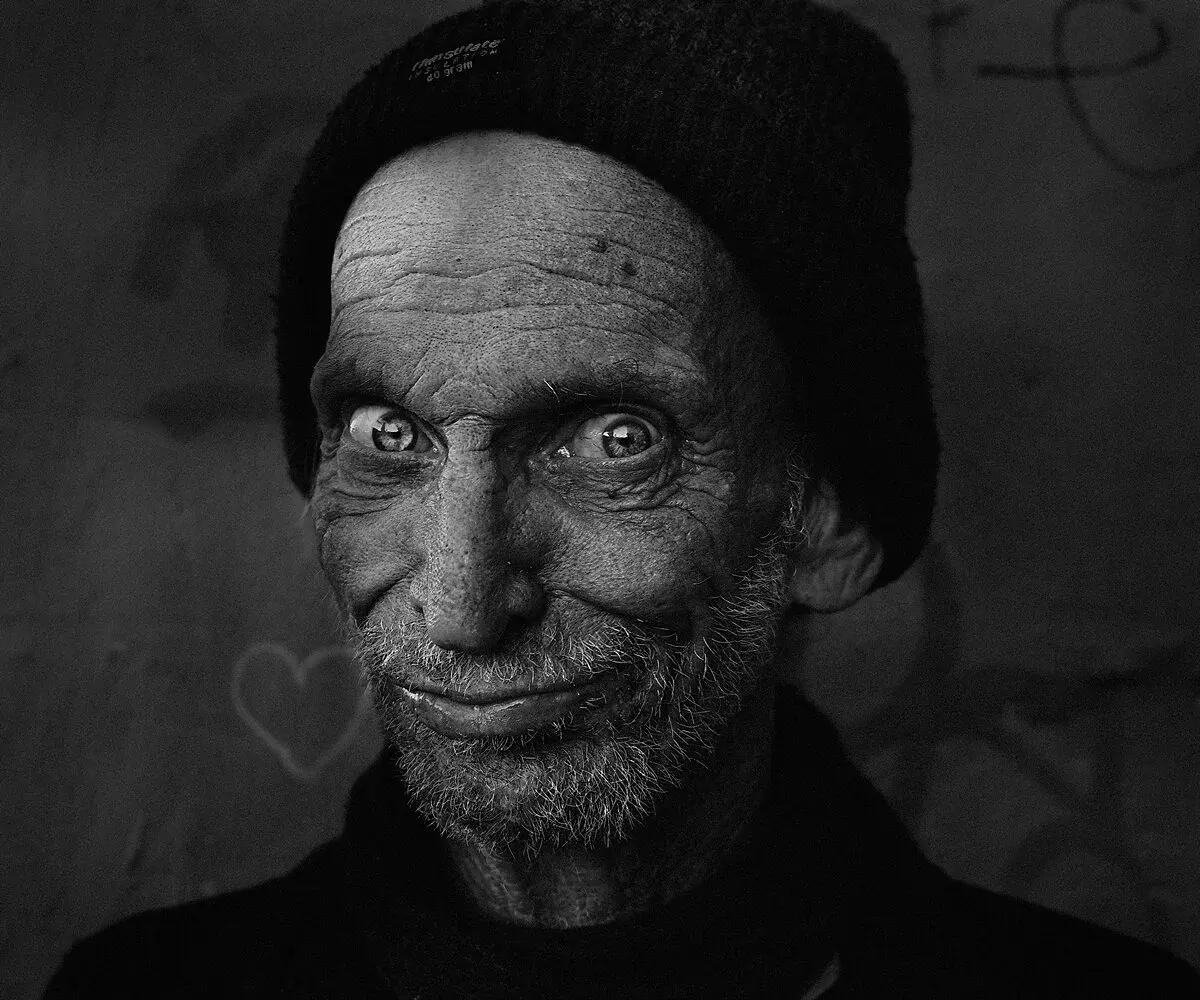 An elderly man in a beanie smiling warmly, a heart drawn in chalk on the wall behind him