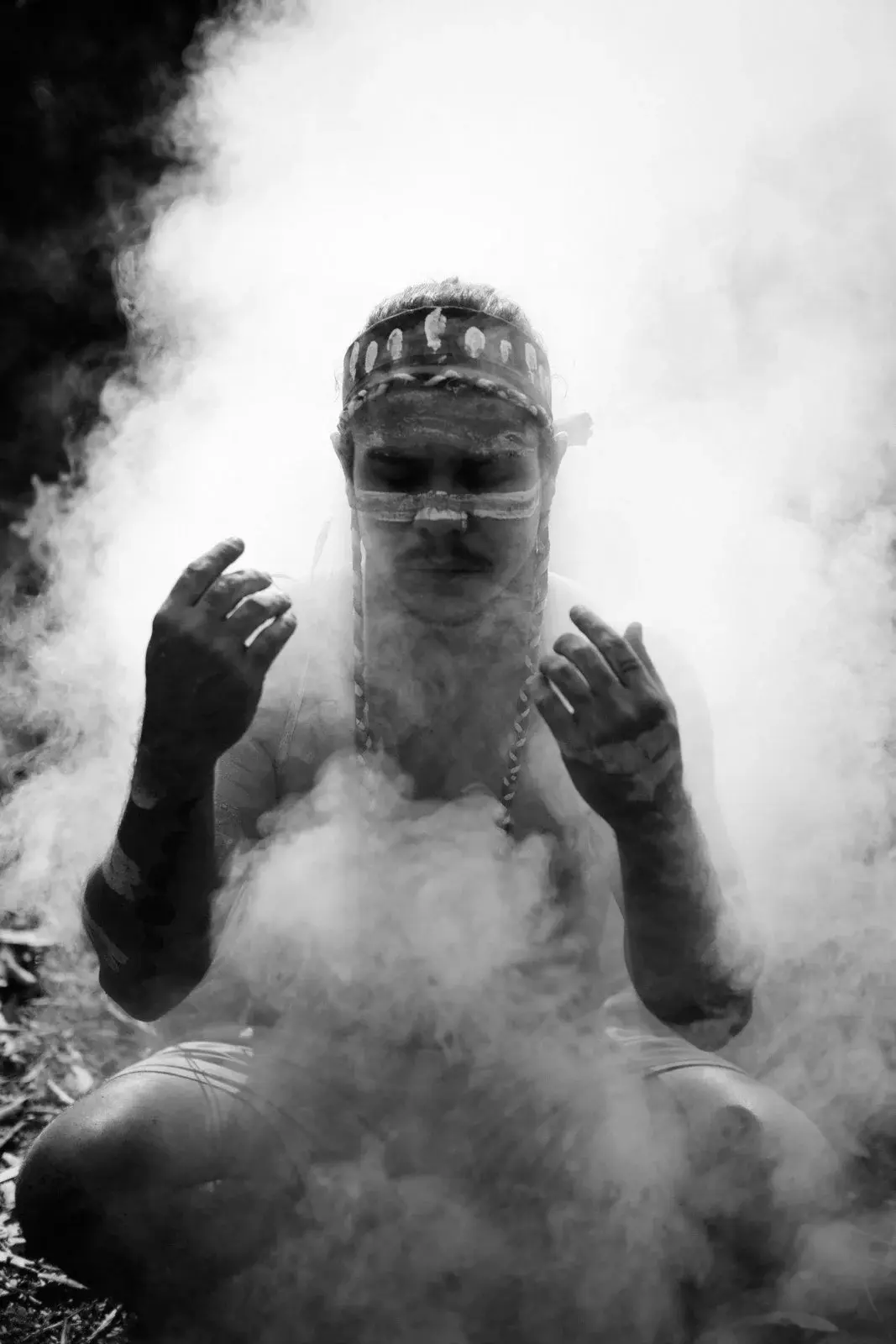 A man crouched in thick white smoke during a ceremony, hands raised, headband marked with ochre