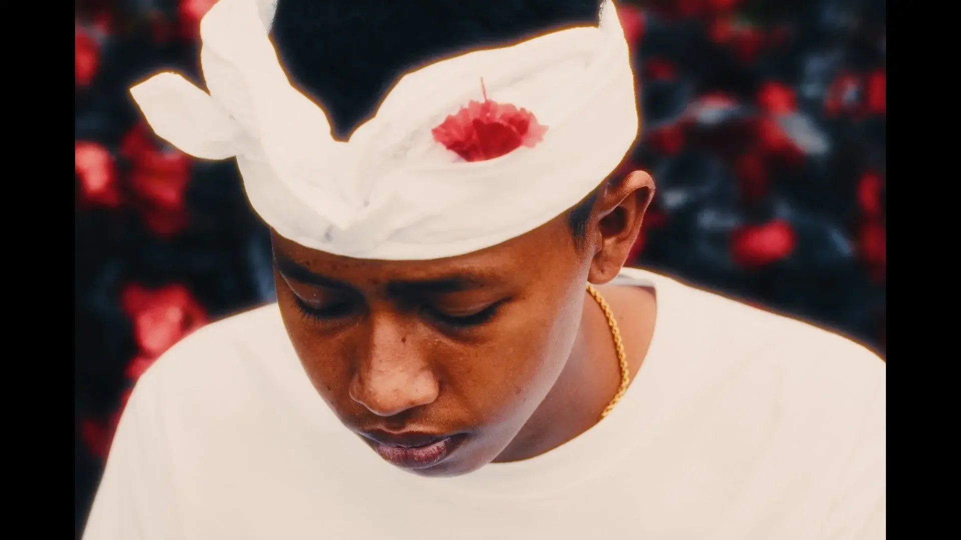 The same boy in white ceremonial headdress with a red flower, looking down