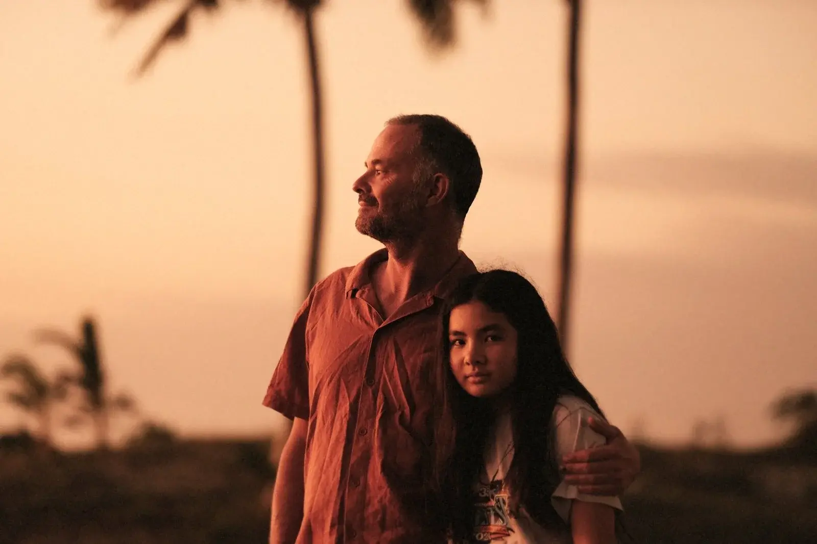 A father with his arm around his daughter at sunset, palm trees behind them