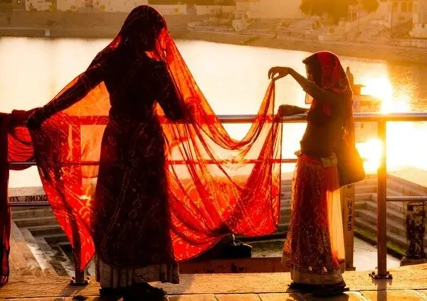 Two women in saris holding a red sari up against the sunset light by the lake at Pushkar