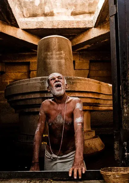 A brahmin priest, ash-marked, calling out before a large stone lingam inside a Shiva temple