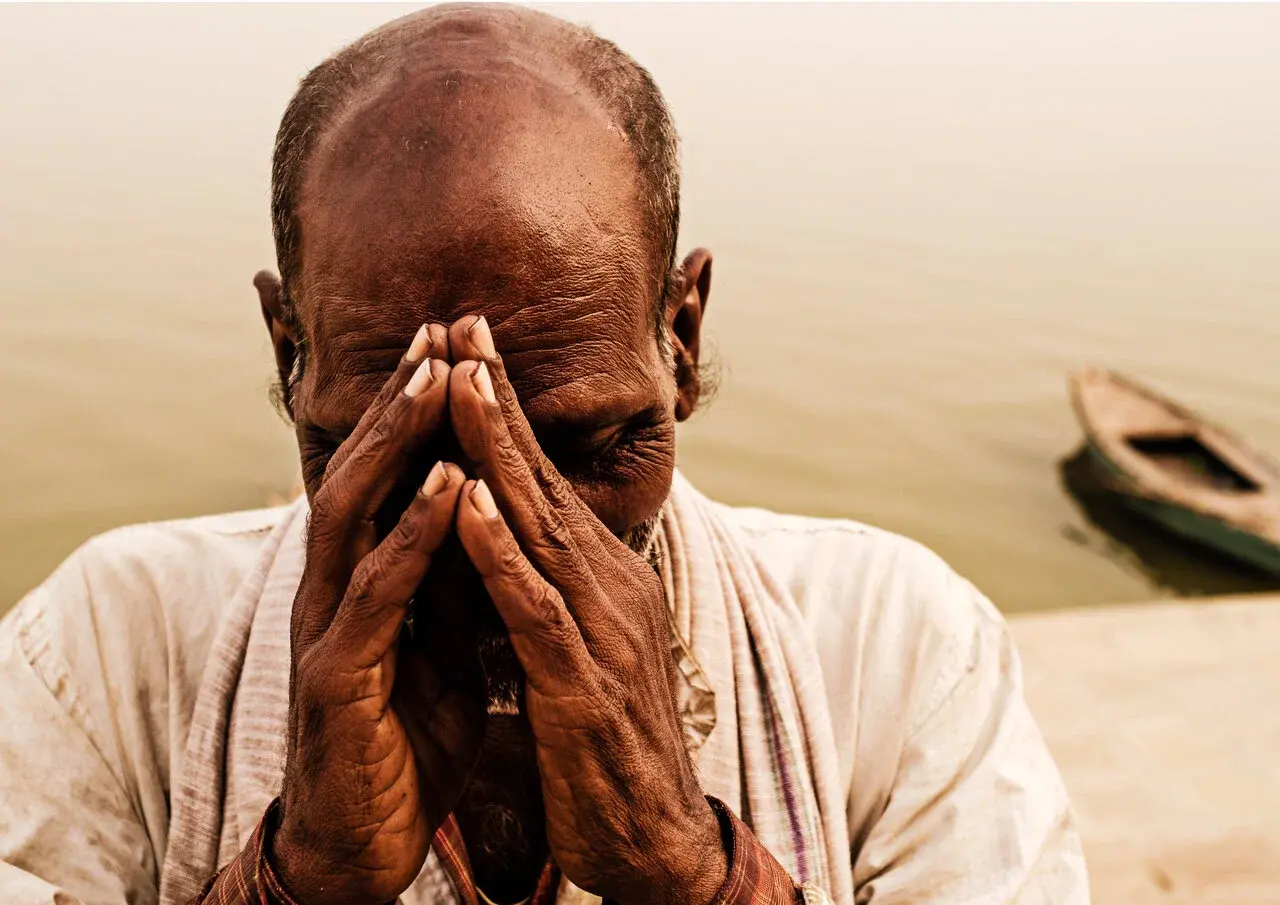 A man praying with his hands pressed together by the Ganges, a boat behind him